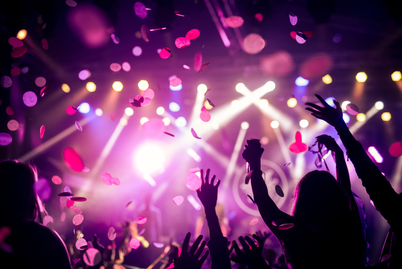 Gente en un concierto con las manos levantadas, confeti cayendo y luces brillantes en el escenario.