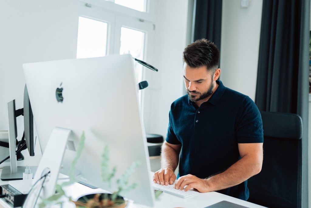 Un hombre tecleando en un escritorio en una oficina moderna y luminosa, junto a un gran monitor iMac.