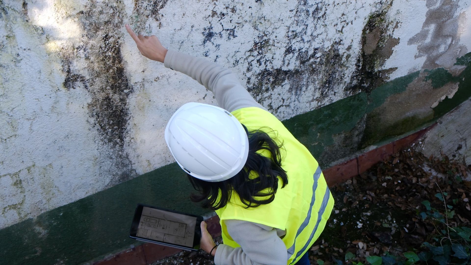 Un ouvrier portant un casque blanc et un gilet jaune analyse un mur taché d'humidité.