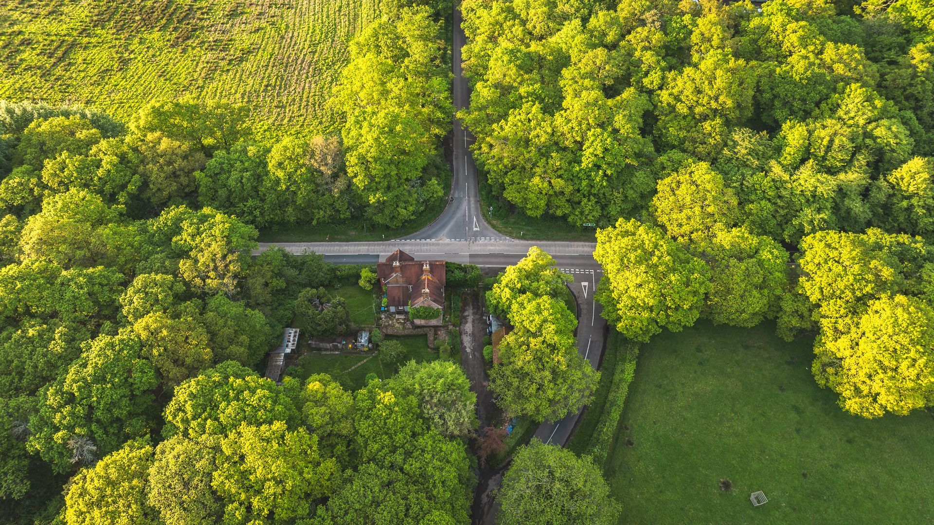 Vue aérienne d'une petite maison en bord de route, entourée d'une forêt dense et verdoyante et de prairies ouvertes.