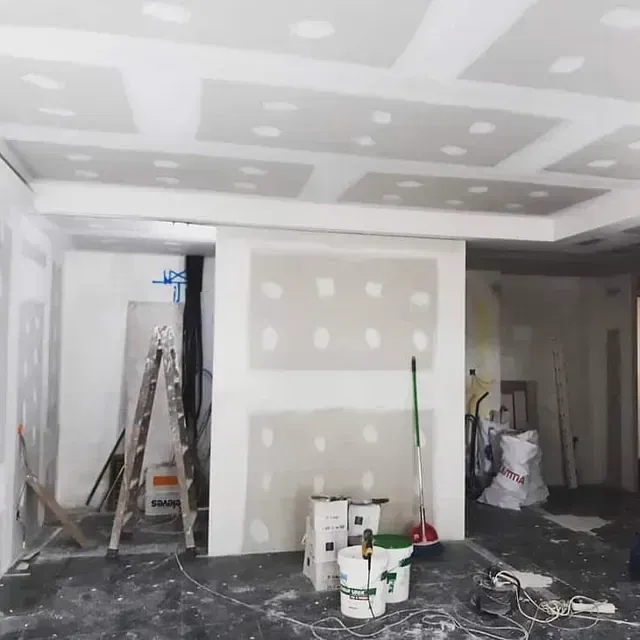 Construcción de interiores: paneles de yeso instalados en paredes y techo, baldes, escalera, escoba y suministros en una habitación.