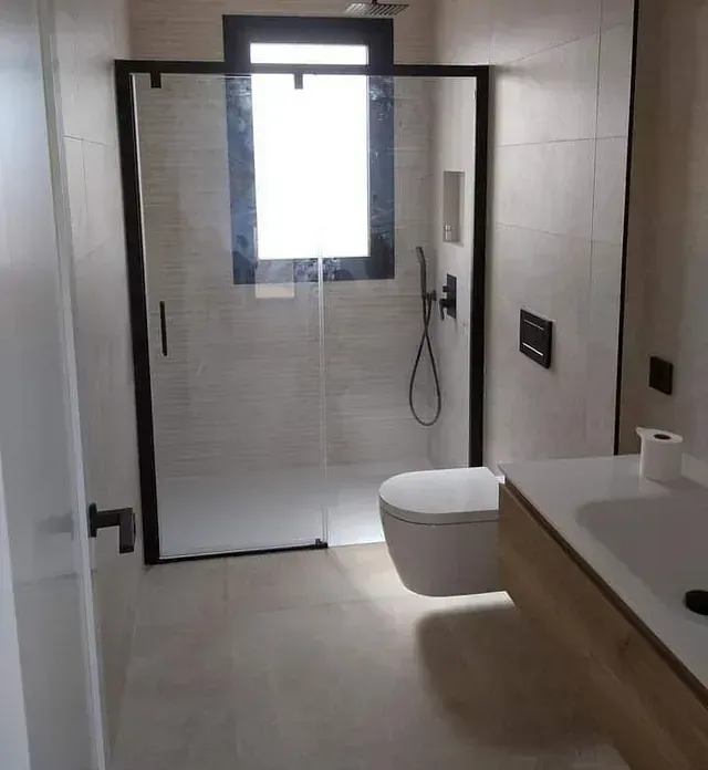 Baño moderno con ducha con marco negro, inodoro y lavabo flotante. Azulejos beige, grifería negra y una pequeña ventana.