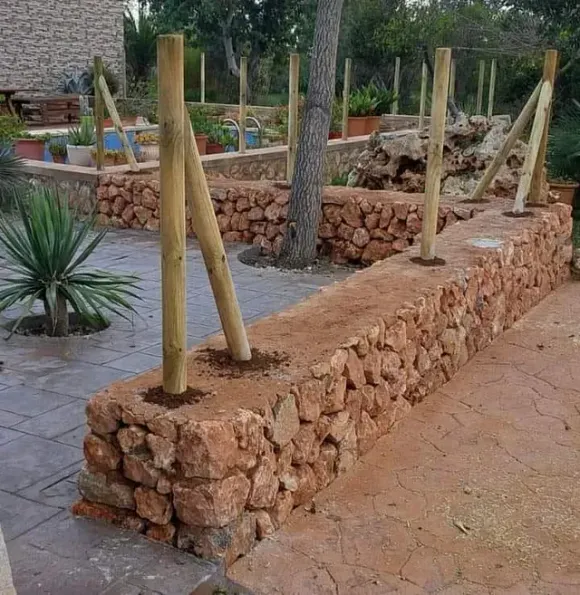 Cama de jardín con paredes de piedra, postes de soporte de madera y un árbol maduro, al aire libre.
