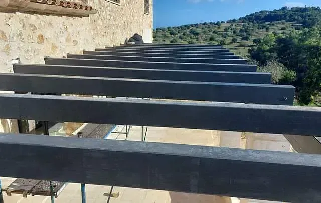 Vigas de pérgola grises sobre un patio, con un edificio de piedra al fondo, bajo un cielo azul.