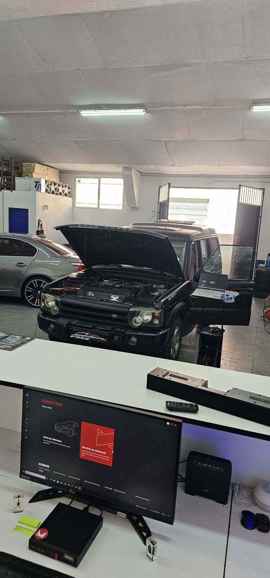 Un coche con el capó levantado está aparcado en un garaje junto a un monitor de ordenador.