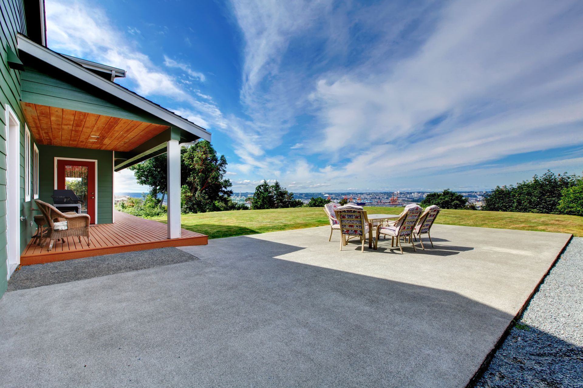 Terrasse avec table et chaises de jardin à côté d'une maison moderne, donnant sur un paysage verdoyant et un ciel bleu.