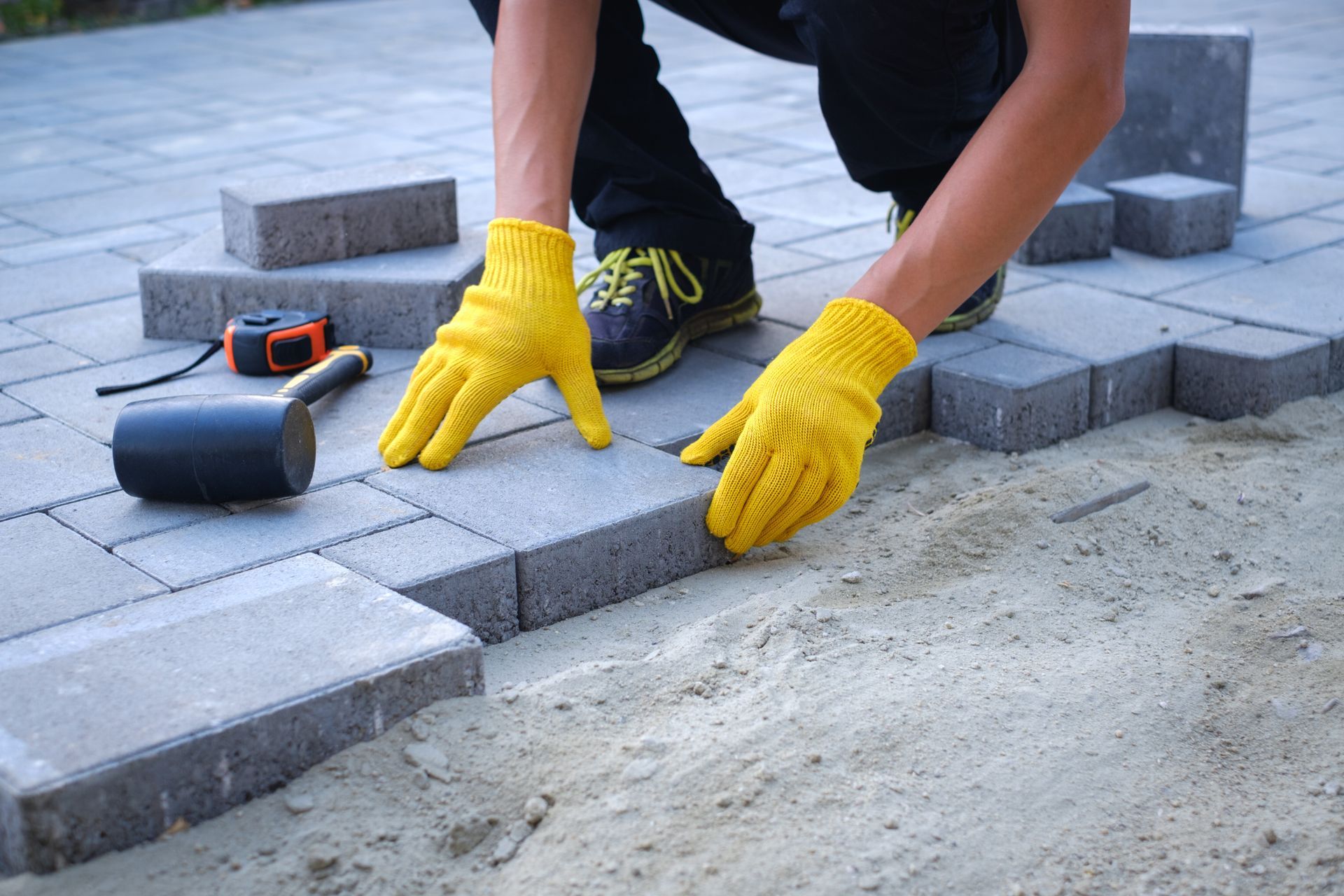 Un ouvrier portant des gants jaunes pose des pavés gris sur une dalle de terrasse.