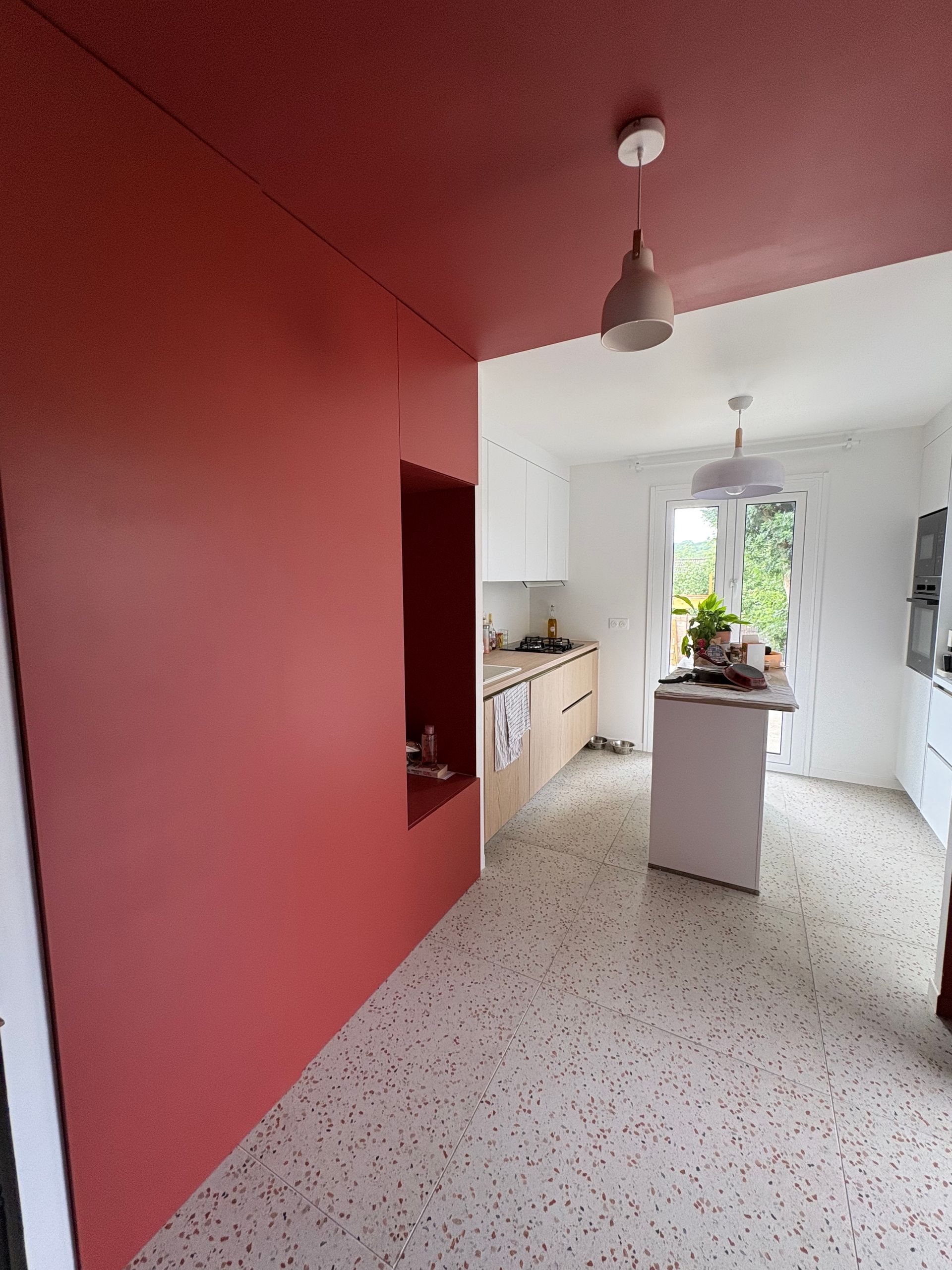 Petite cuisine avec un mur rouge, des placards blancs, un sol en terrazzo et des plantes sur le rebord de la fenêtre.