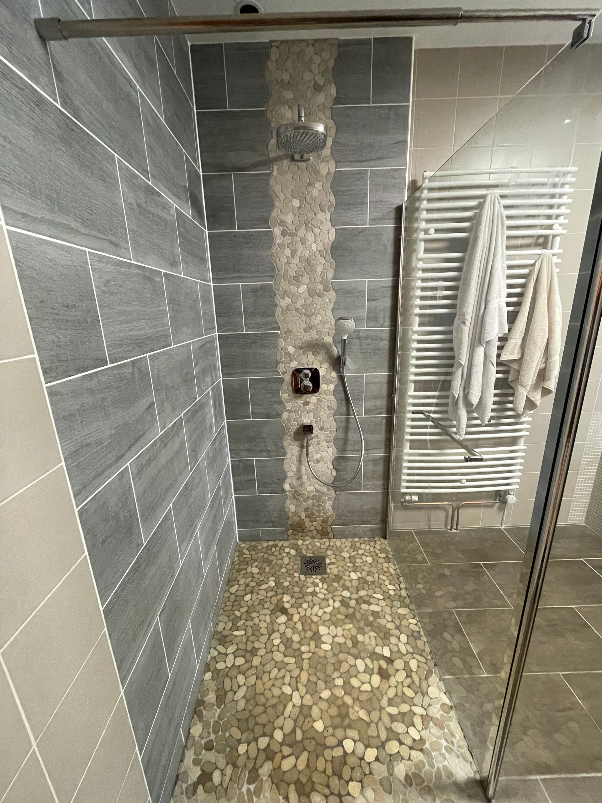 Petite salle de bains avec murs carrelés gris, sol en galets, espace douche et sèche-serviettes blanc.