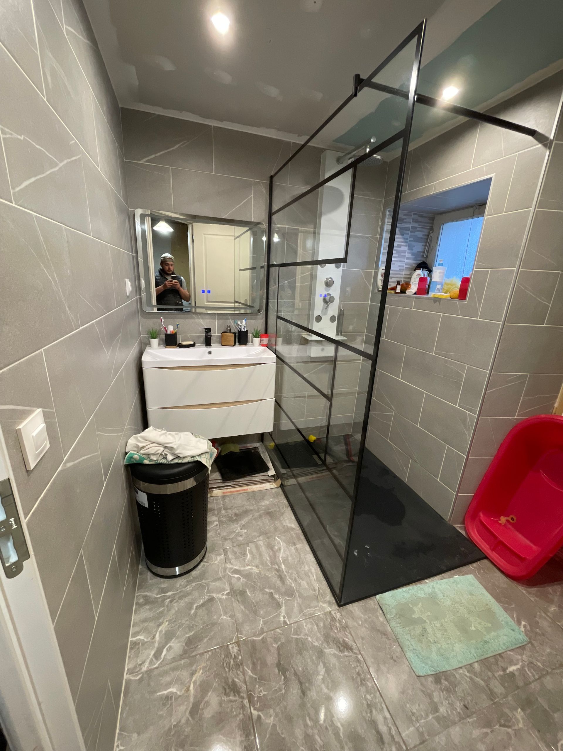 Salle de bains moderne carrelée de gris avec lavabo, miroir, douche en verre, poubelle et chaise rouge dans le coin