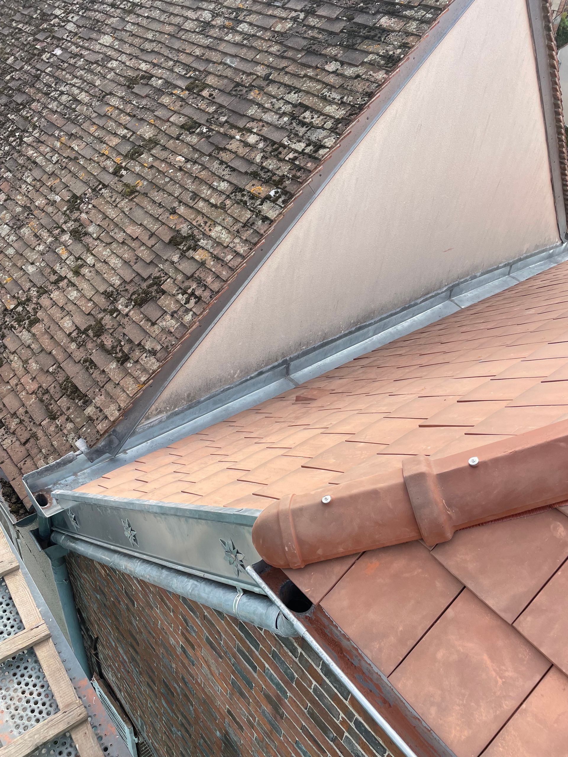 Vue aérienne d'une gouttière et de pans de toiture en tuiles entrecroisés, avec un mur beige et des tuiles brunes.