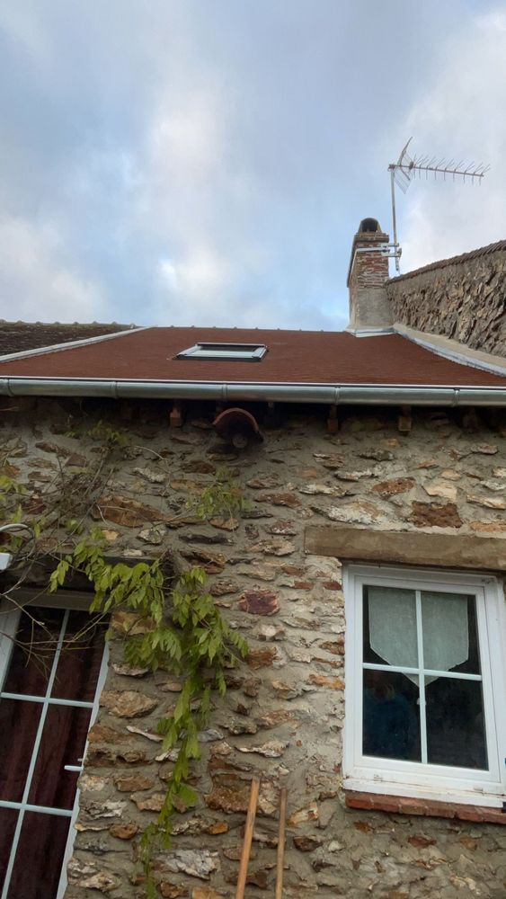 Extérieur d'une maison en pierre avec toit rouge, cheminée, fenêtre à cadre blanc et antenne de télévision sur fond de ciel nuageux
