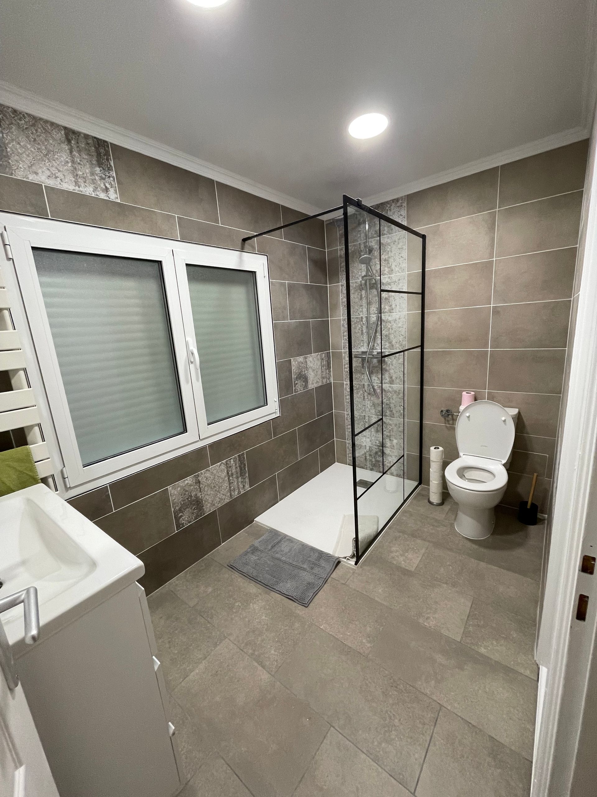 Salle de bains moderne avec murs carrelés, douche en verre, toilettes, fenêtre et meuble-lavabo