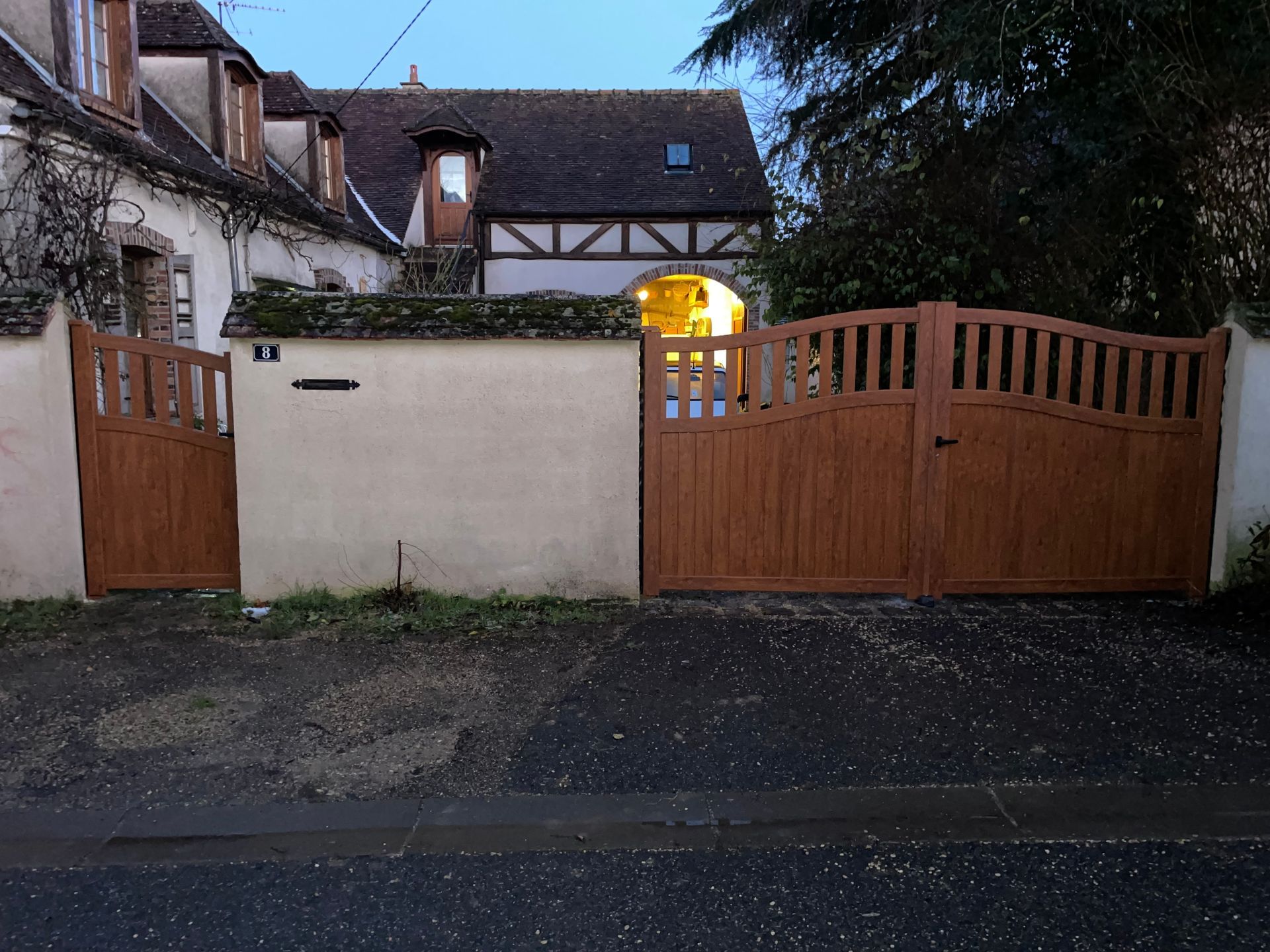 Portail en bois et mur couleur crème à l'entrée d'une maison au crépuscule, avec une fenêtre éclairée derrière.