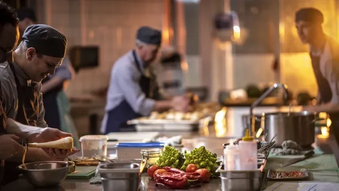 Köche bereiten in einer geschäftigen Großküche Speisen zu. Gemüse und Zutaten liegen auf einer Arbeitsfläche.