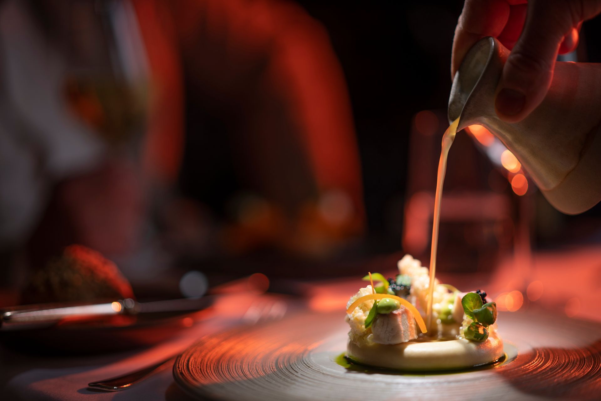 Een hand giet saus op een gastronomisch gerecht op een donker, roodgloeiend oppervlak.