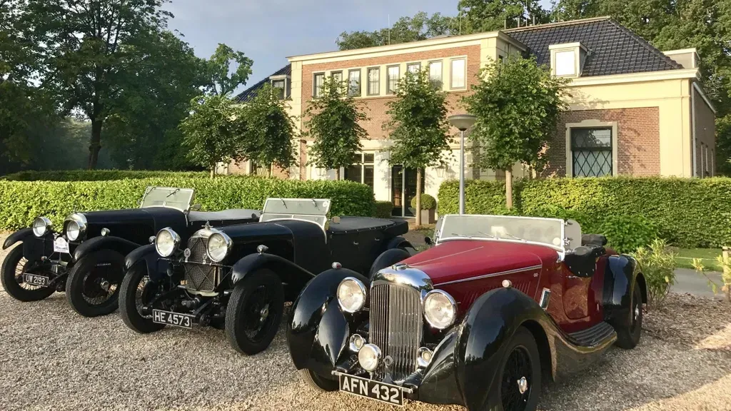 Vier klassieke zwart-rode auto's geparkeerd voor een bakstenen gebouw met bomen.