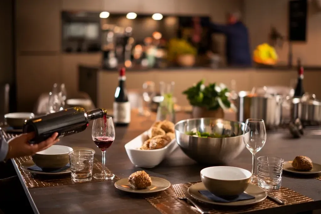 Een persoon schenkt rode wijn in een glas terwijl hij aan de eettafel staat, die gedekt is voor een maaltijd.