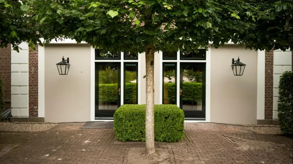 Een boomstam in het midden van een gebouw met ramen, waarin groen weerspiegeld is, geflankeerd door lantaarns en metselwerk.