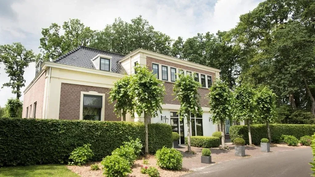 Bakstenen huis met twee verdiepingen, verzorgde tuin, bomen en een oprit van donker asfalt.