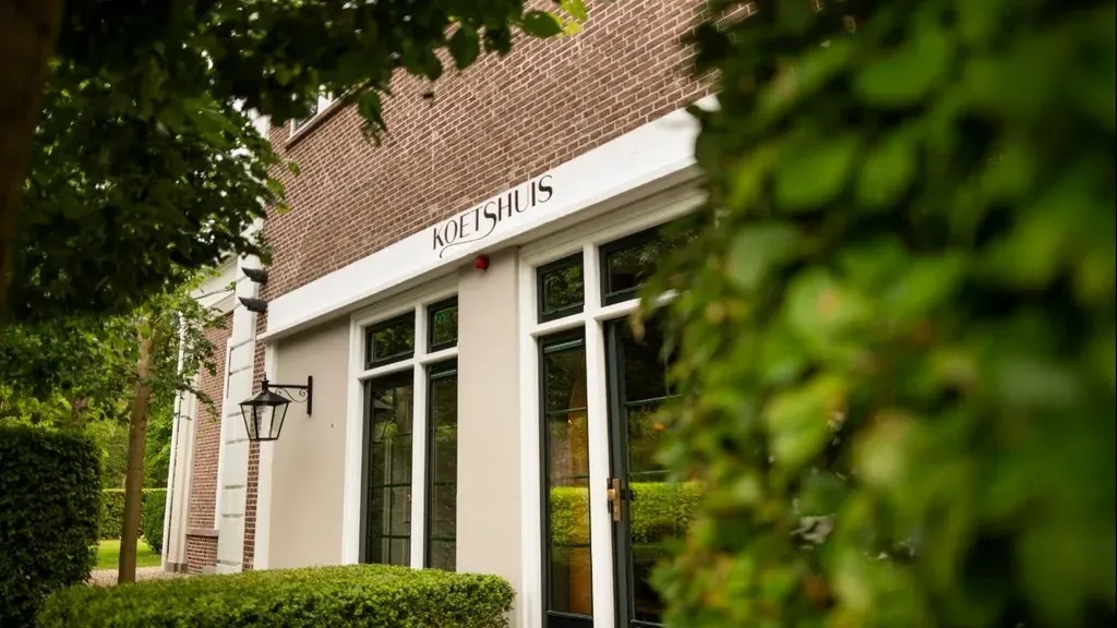 Buitenkant van restaurantgebouw met witte afwerking, bruine bakstenen gevel en groen gebladerte.