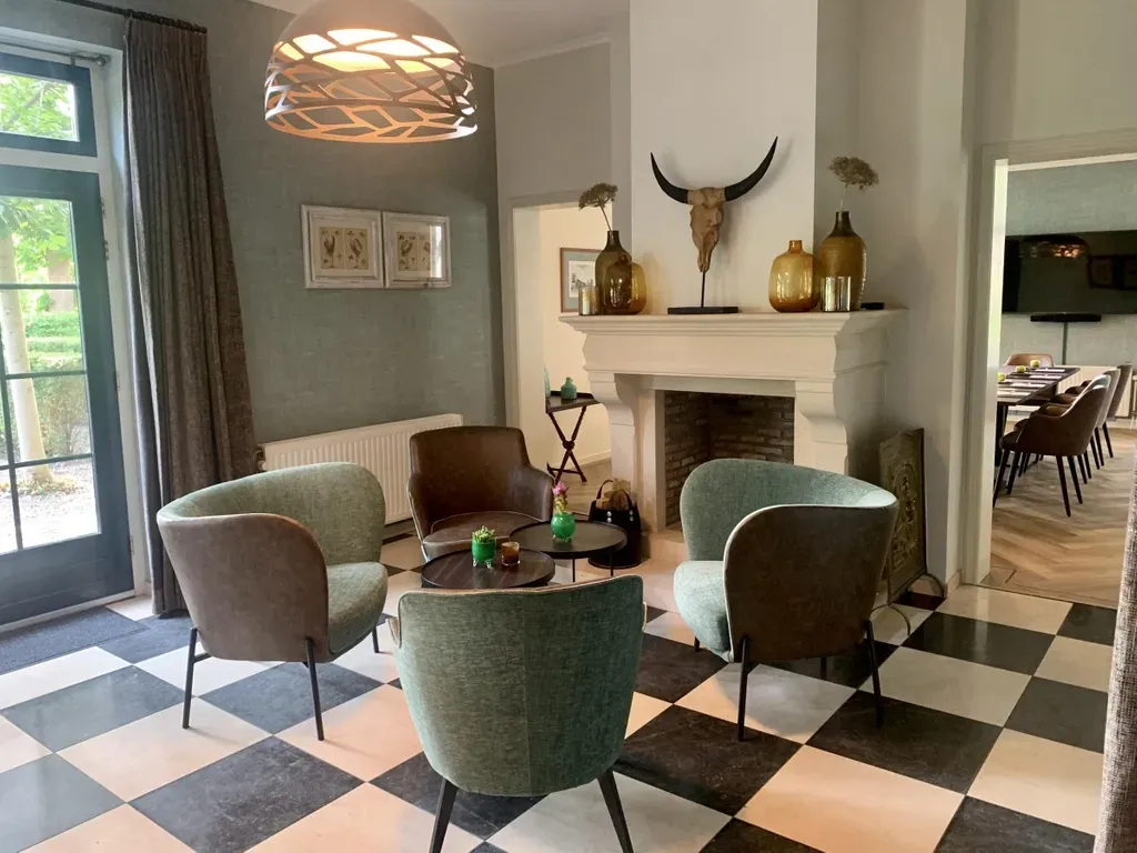 Woonkamer met zithoek, zwart-wit geblokte vloer, open haard en groene stoelen.