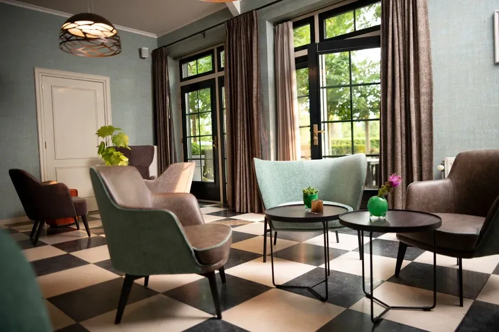 Loungegedeelte met fauteuils rond kleine tafeltjes op een geblokte vloer, grote ramen en gordijnen met patroontjes.