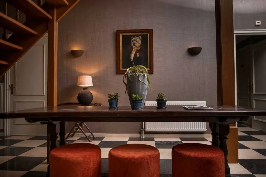 Elegante entree met een lange houten tafel, rode poefs en kunstwerken. Een trap en een geblokte vloer zijn zichtbaar.