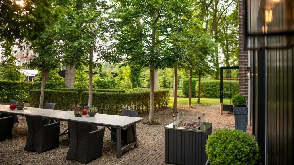 Eetgedeelte buiten met lange tafel, bomen en verzorgde tuin.