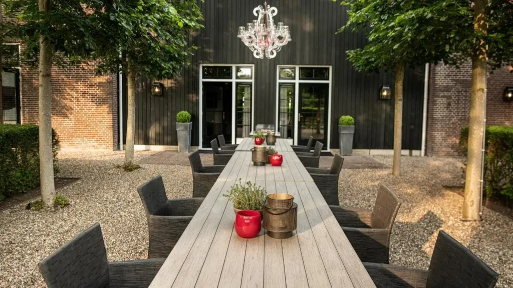 Eetgedeelte in de openlucht met een lange tafel, stoelen en bomen, voor een donker gebouw met een kroonluchter.