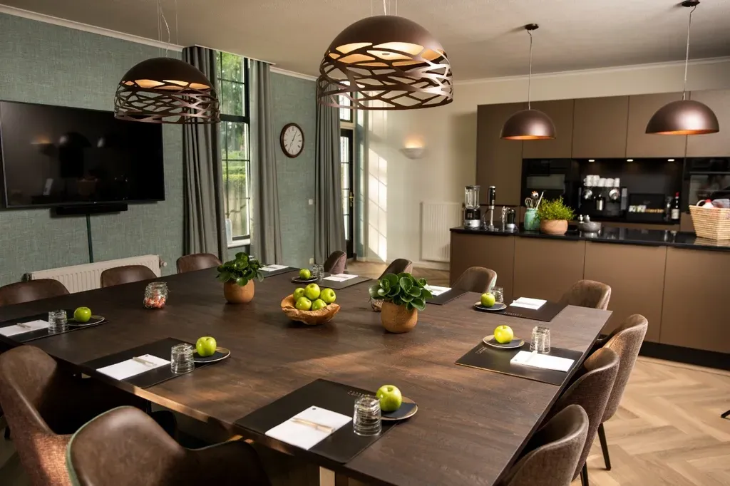 Vergaderruimte met grote houten tafel, bruine stoelen en keukenhoek. Groene muur, donkere plafondlampen.