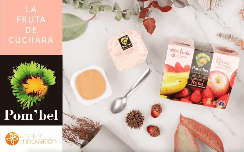 Envase del postre de frutas Pom'bel con un tazón de puré de fruta, una cuchara y frutos secos y hojas decorativas sobre una superficie blanca.