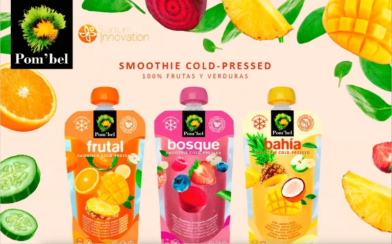Tres bolsitas de batido de frutas y verduras prensado en frío de la marca Pom'bel, en colores naranja, rosa y amarillo, rodeadas de productos frescos.