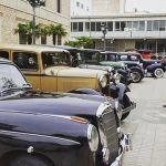 Coches antiguos aparcados frente a un edificio, con un taxi beige y gente reunida cerca de la entrada.