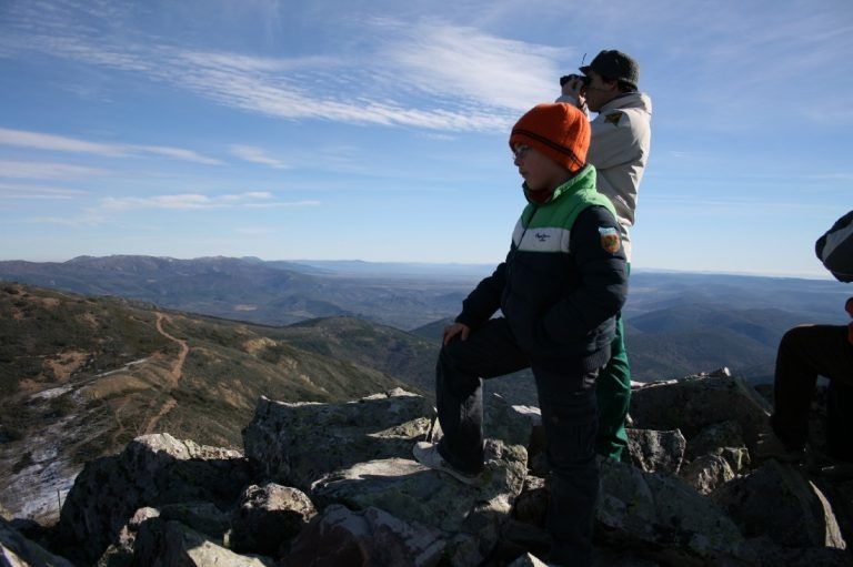 Dos excursionistas con ropa de invierno contemplan un paisaje montañoso rocoso bajo un cielo azul.