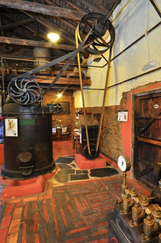 Antigua sala de calderas industrial con una gran máquina negra, tuberías, manómetros y paredes de ladrillo.
