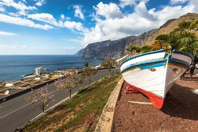 Un barco blanco y azul en exhibición con vista a una carretera costera, edificios y el océano con acantilados en el fondo.