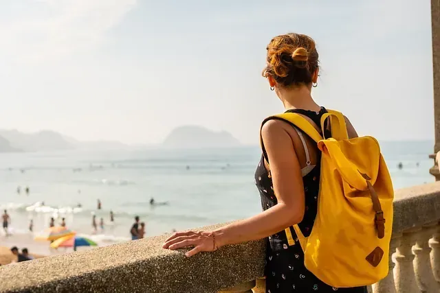 Mujer con una mochila amarilla mirando una playa y el océano desde un balcón.