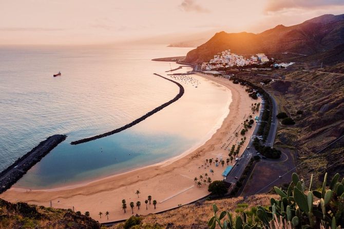 Escena de playa al atardecer. La arena dorada se curva a lo largo de la costa con un rompeolas de roca. Montañas y edificios al fondo.