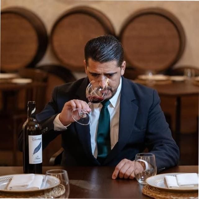 Hombre de traje oliendo vino tinto, botella de vino y copas sobre una mesa, barriles al fondo.