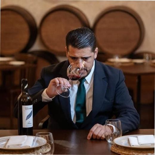 Hombre de traje oliendo vino tinto, botella de vino y copas sobre una mesa, barriles al fondo.