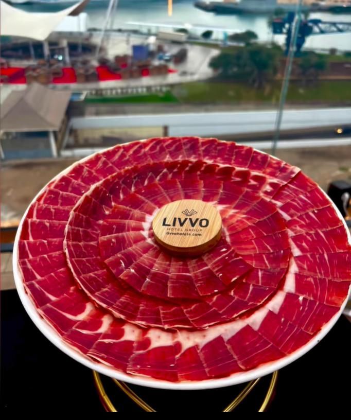 Plato de jamón curado en lonchas finas, dispuesto en círculos concéntricos.  Vista de ventana al fondo.