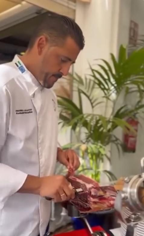 Chef cortando jamón curado a cuchillo, con expresión concentrada. Restaurante con plantas verdes.