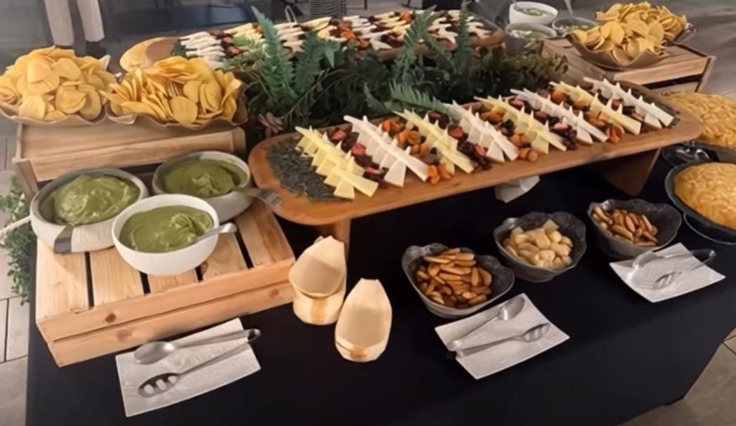 Buffet con patatas fritas, guacamole, queso, frutos secos y aperitivos dispuestos sobre una mesa y expositores de madera.