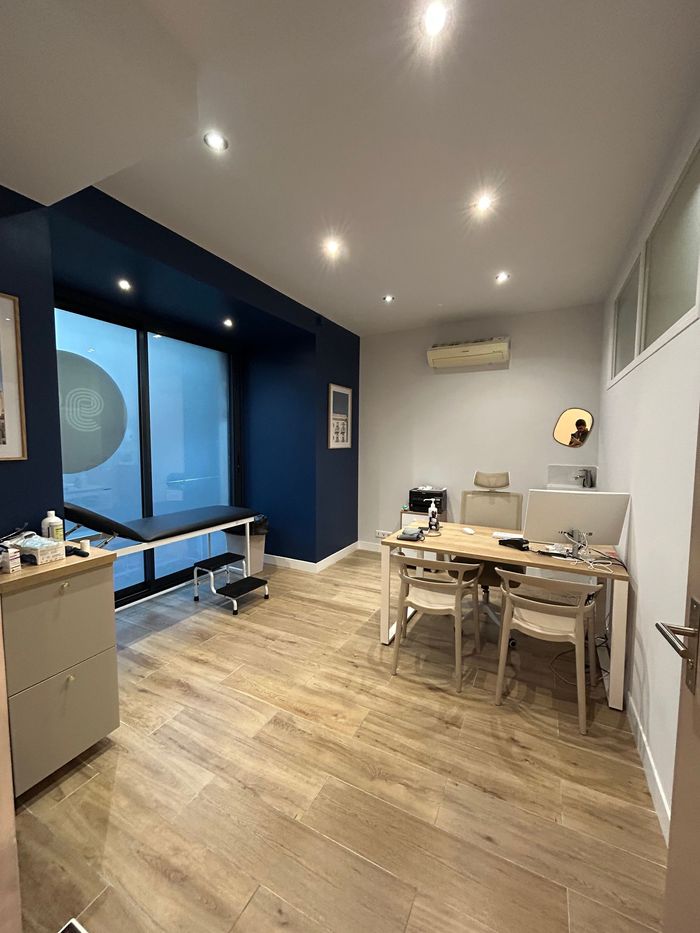 Salle d'examen médical : table d'examen, table avec chaises, mur bleu foncé, parquet clair.