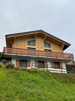 Chalet en bois avec balcon, situé à flanc de colline herbeuse sous un ciel nuageux.
