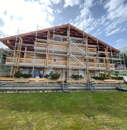 Des échafaudages entourent un bâtiment en bois à plusieurs étages, situé sur une pelouse sous un ciel nuageux.