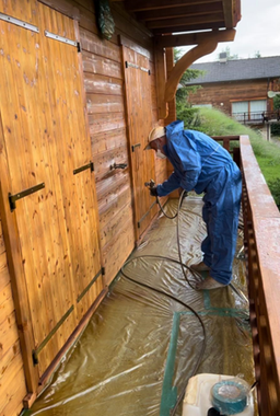 Une personne en combinaison de protection bleue vaporise des portes en bois à l'extérieur.