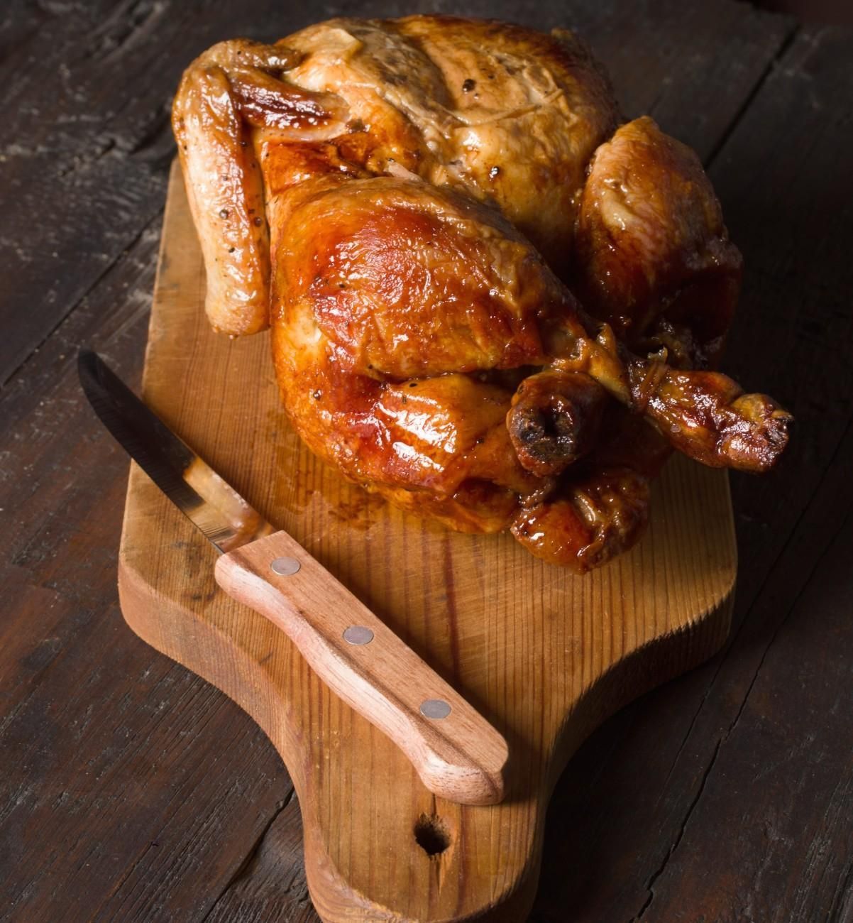 Un pollo asado sobre una tabla de cortar de madera con un cuchillo al lado, sobre un fondo rústico oscuro.