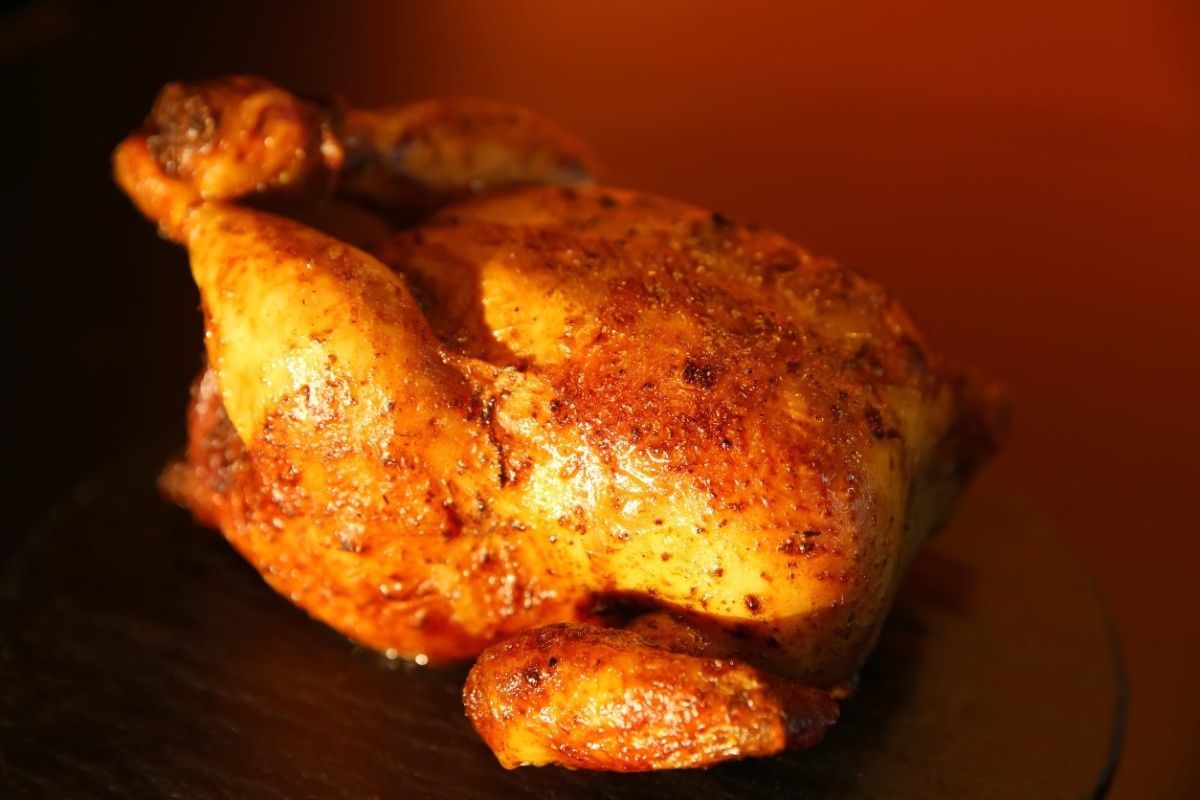 Un pollo entero asado de color marrón dorado, centrado sobre una superficie oscura con un fondo cálido de tono anaranjado.